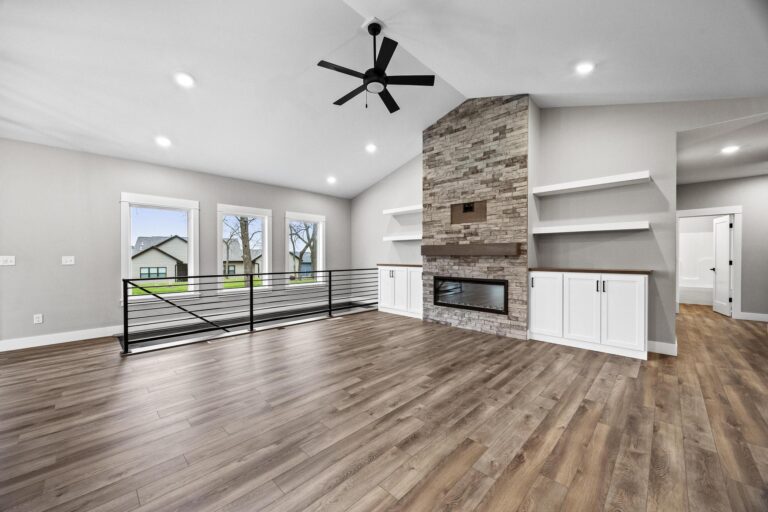 living room with vaulted ceiling, ceiling fan, brick gas firepalce, metal railing on stairs; large windows to backyard